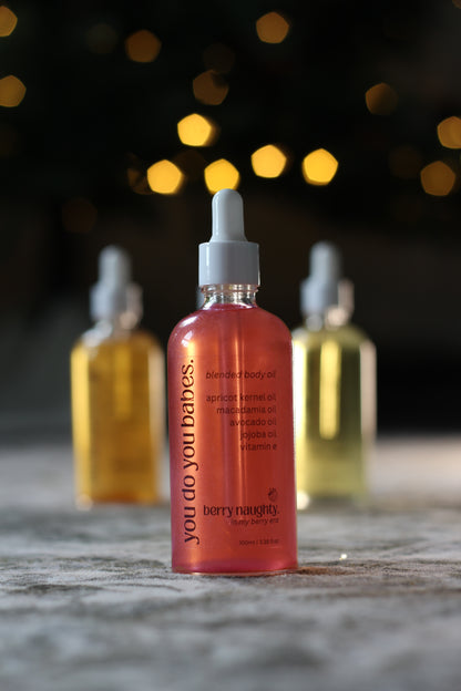 Bottle of 'you do you babes' berry naughty body oil on a wooden surface with blurred lights in the background.