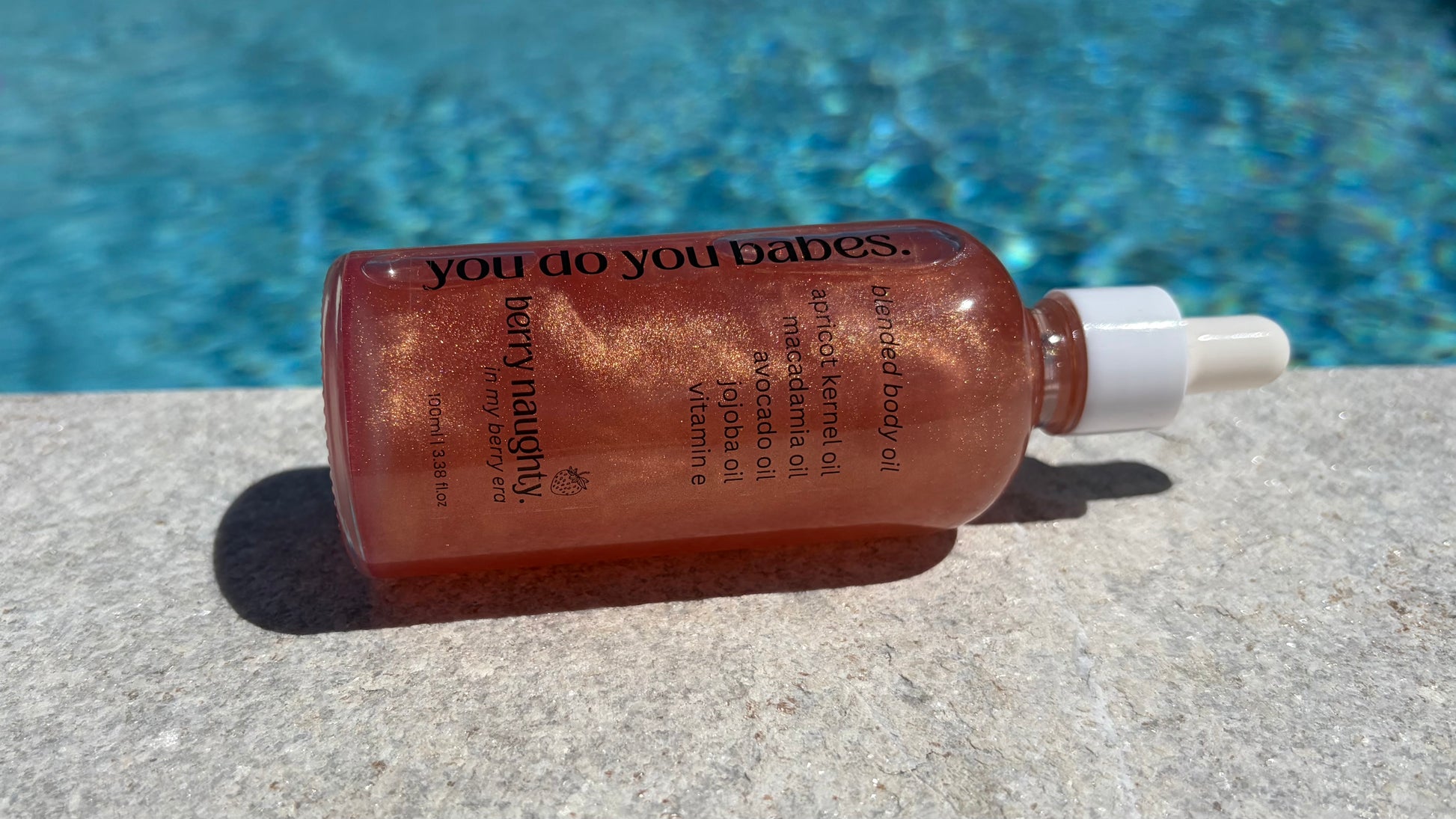 Bottle of 'you do your babes' body oil berry naughty product on a concrete surface with a pool in the background