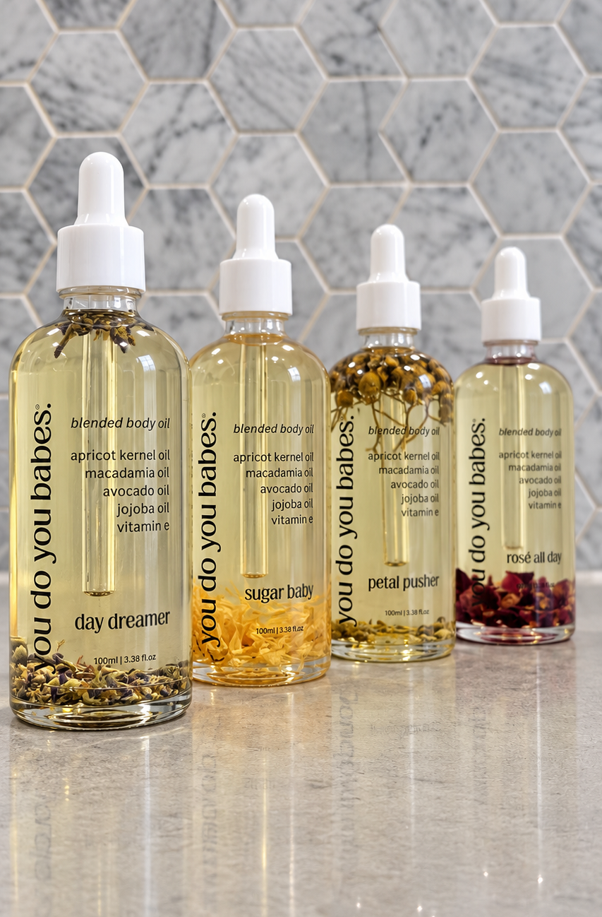 Four clear dropper bottles of body oil arranged on a glossy surface against a gray hexagonal tile wall. The bottles contain pale yellow oil with visible botanical pieces at the bottom, and the labels read 'you do you babes.