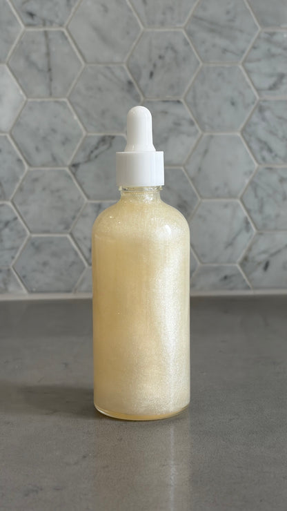 Bottle of 'you do you babes' coco glow blended body oil against a grey tiled wall.