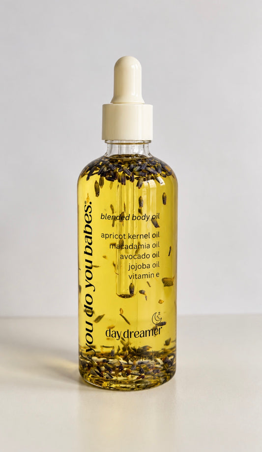 A clear glass dropper bottle filled with golden yellow body oil and containing visible dried botanical pieces, with a cream-colored dropper cap. The bottle is labeled 'you do you babes: blended body oil' and shows the brand 'daydreamer'.