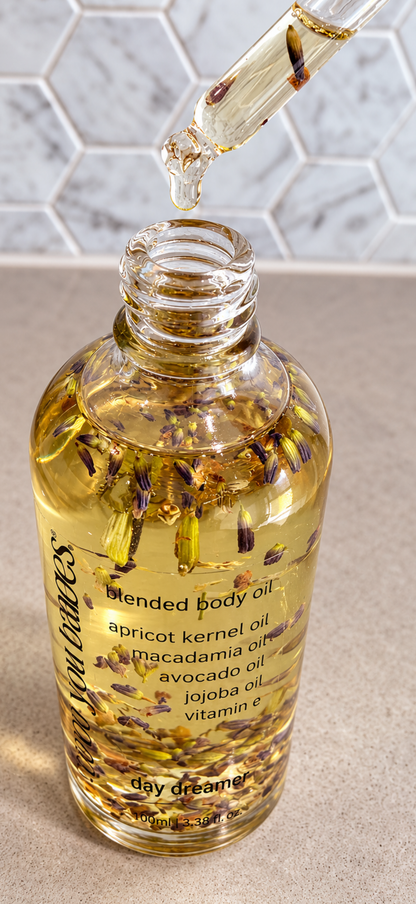 A clear glass bottle of Day Dreamers blended body oil filled with golden yellow oil and suspended botanical pieces, with a glass dropper above the open bottle. The background shows a light gray marble tile wall and a beige countertop.