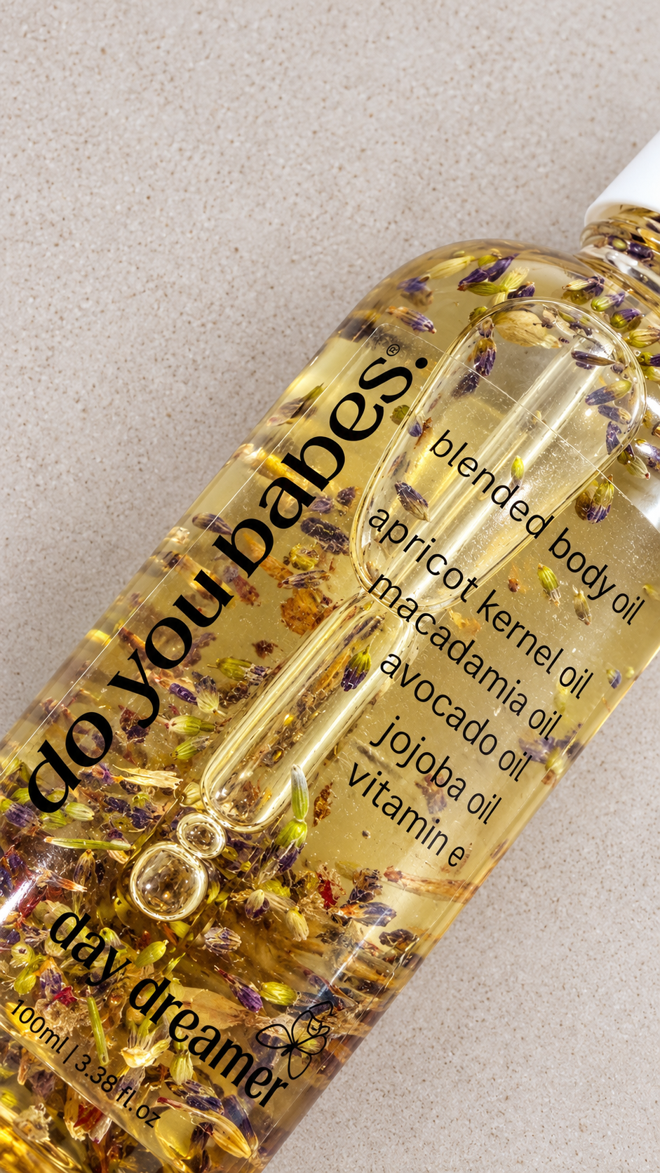 A close-up of a clear bottle of 'do you babes.' Day Dreamer blended body oil, filled with golden oil and suspended dried botanicals, on a light textured background.