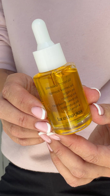 A person holding a small glass dropper bottle filled with golden yellow serum or oil. The bottle has a white dropper cap and is called face the glow facial oil 