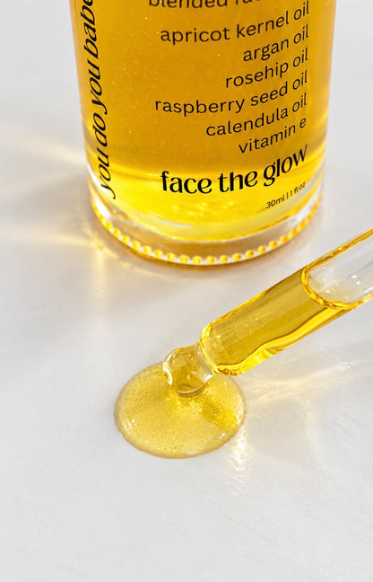 A close-up of a clear glass bottle filled with golden yellow facial oil, with a glass pipette resting on a white surface and a small pool of the oil in front. The bottle label shows the brand name 'face the glow' by you do you babes and lists ingredients including apricot kernel oil, argan oil, rosehip oil, raspberry seed oil, and calendula oil.