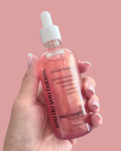 Hand holding a pink bottle of Berry Naughty 'you do you babes' body oil against a pink background