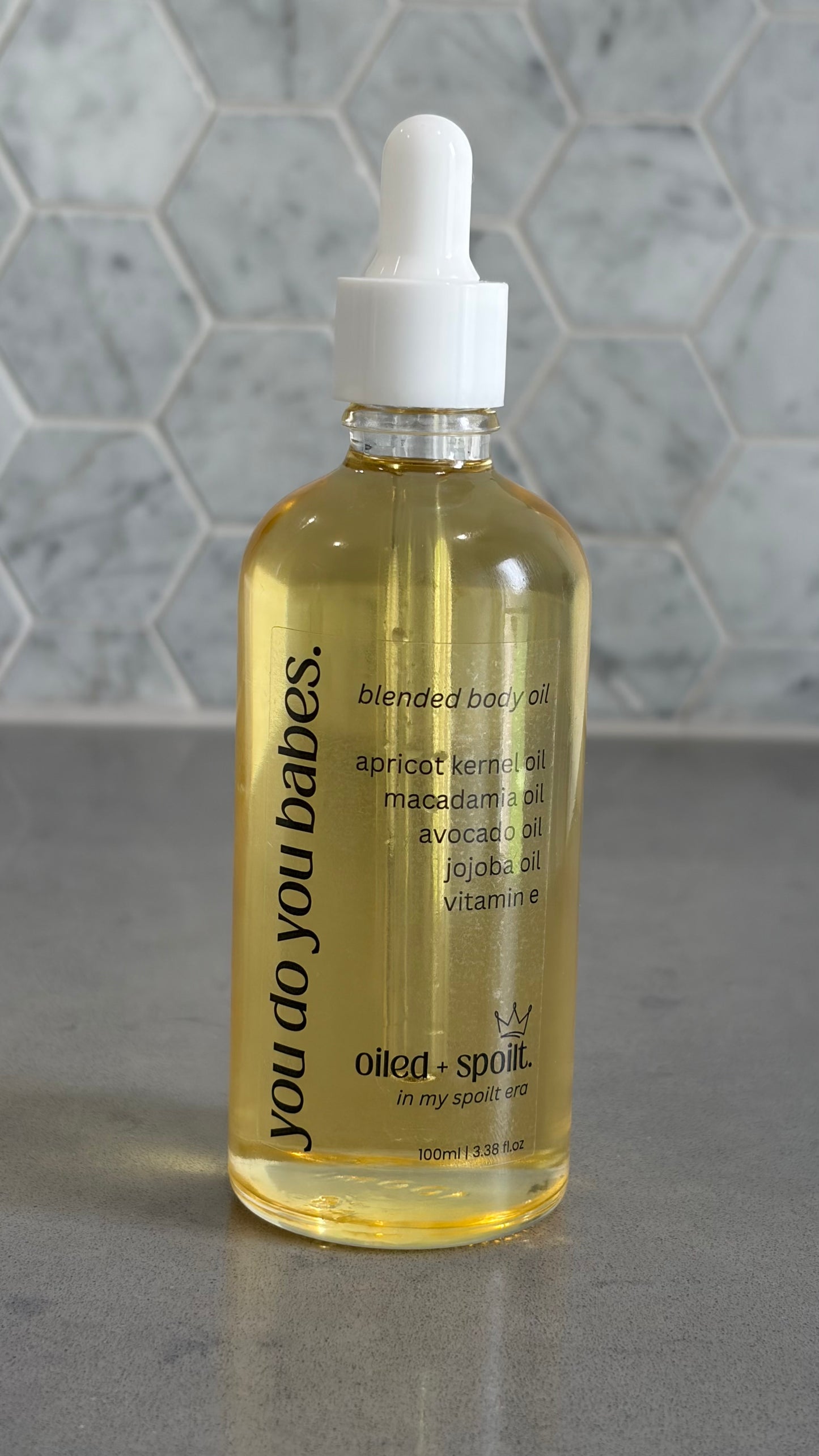 Bottle of 'you do you babes' blended body oil with a dropper on a gray surface.