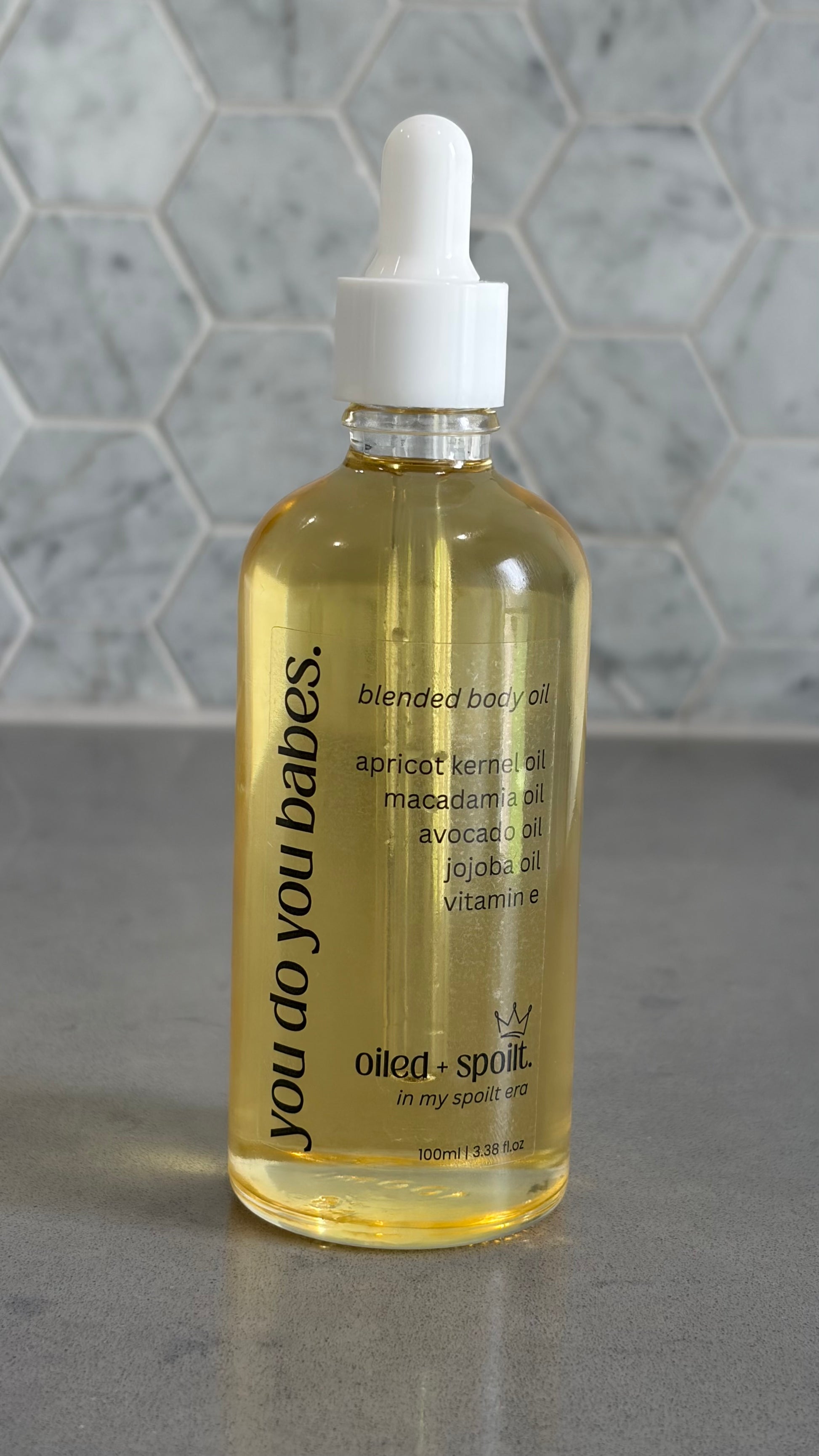 Bottle of 'you do you babes' blended body oil with a dropper on a gray surface.