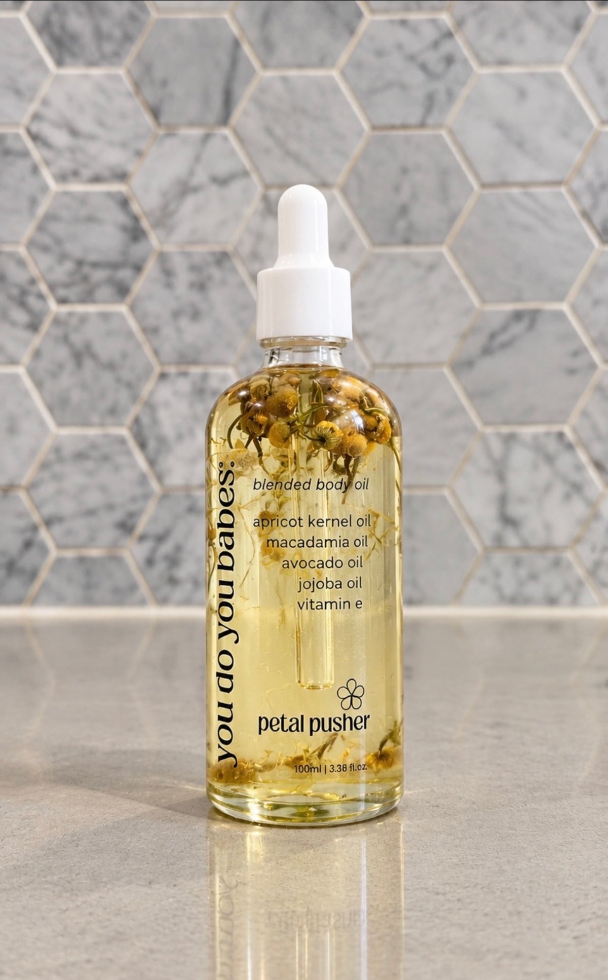 A bottle of Petal Pusher 'you do you babes!' enriched body oil with a white dropper cap, filled with golden oil and botanicals, set against a gray marble hexagonal tile background.