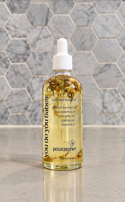A bottle of Petal Pusher 'you do you babes!' enriched body oil with a white dropper cap, filled with golden oil and botanicals, set against a gray marble hexagonal tile background.