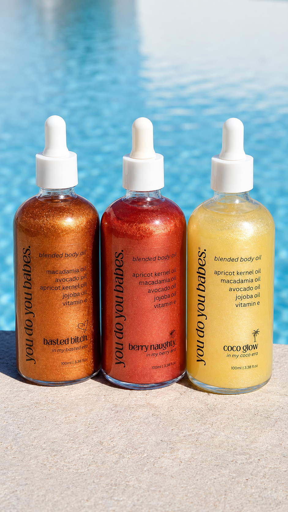 Three dropper bottles of body oil arranged on a light surface with a blurred blue pool in the background. The bottles contain shimmering oils