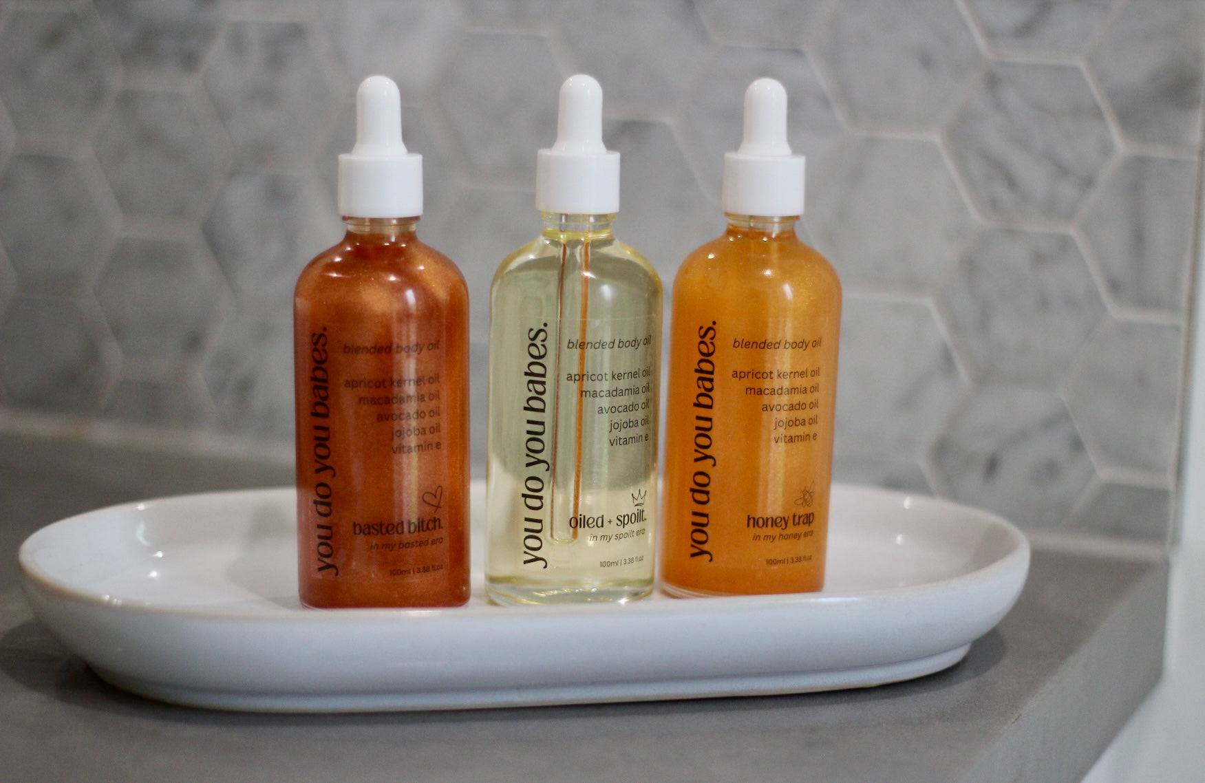 Three bottles of 'You Go You Babe' body oils on a white tray with a gray tiled background.