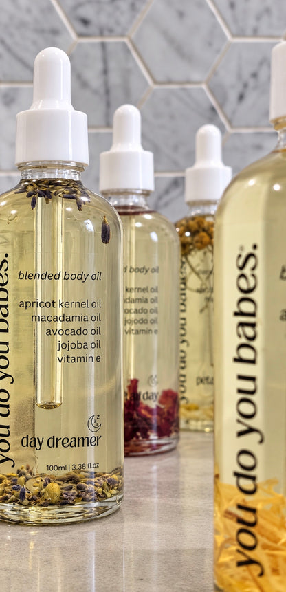 Four clear dropper bottles of Day Dreamer body oils arranged on a countertop against a gray hexagonal tile backsplash. The front bottle is fully visible and labeled 'blended body oil' with botanicals inside; the other bottles are partially cropped in the background and foreground.