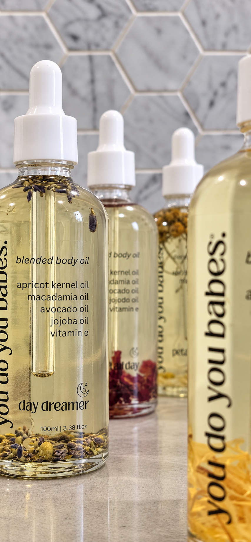 A row of clear glass dropper bottles of Day Dreamer blended body oil with white droppers, arranged on a countertop against a gray hexagonal tile backsplash. The bottles contain golden-yellow oil with visible botanical ingredients at the bottom.