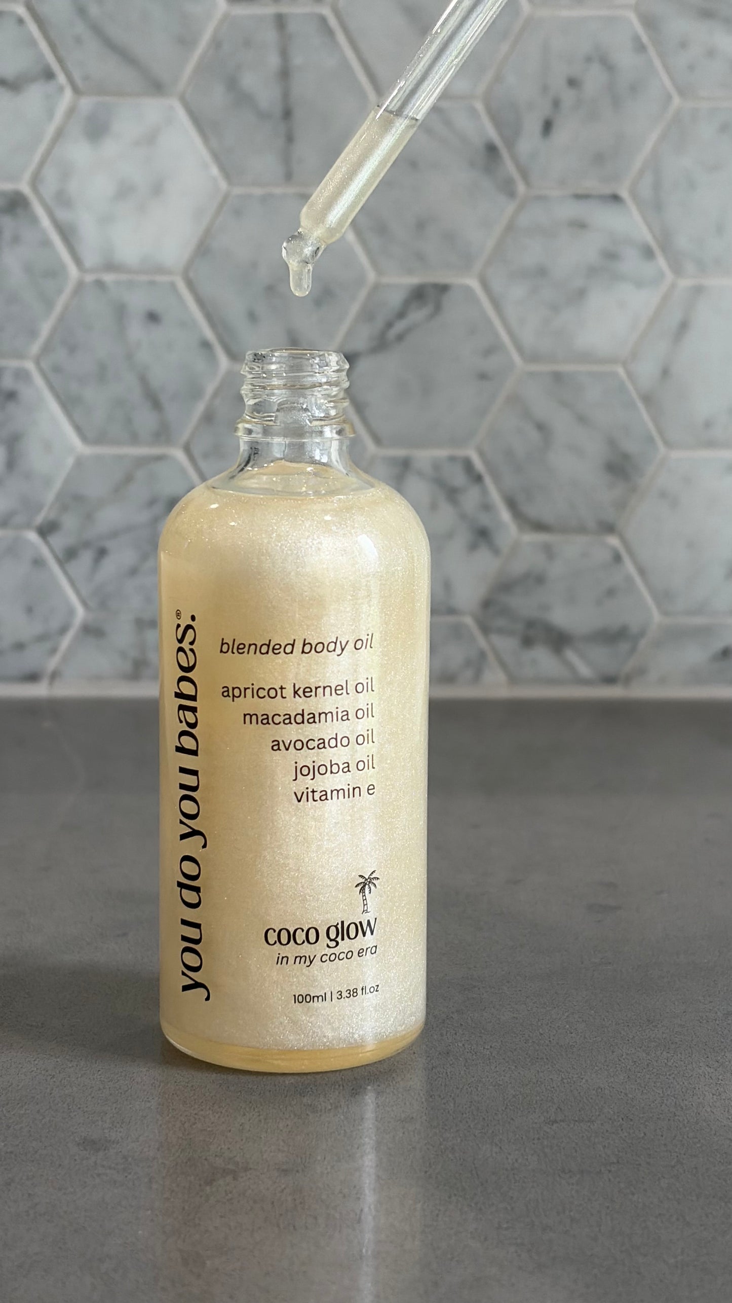 Bottle of 'you do you babes' coco glow blended body oil with a dropper on a grey surface.