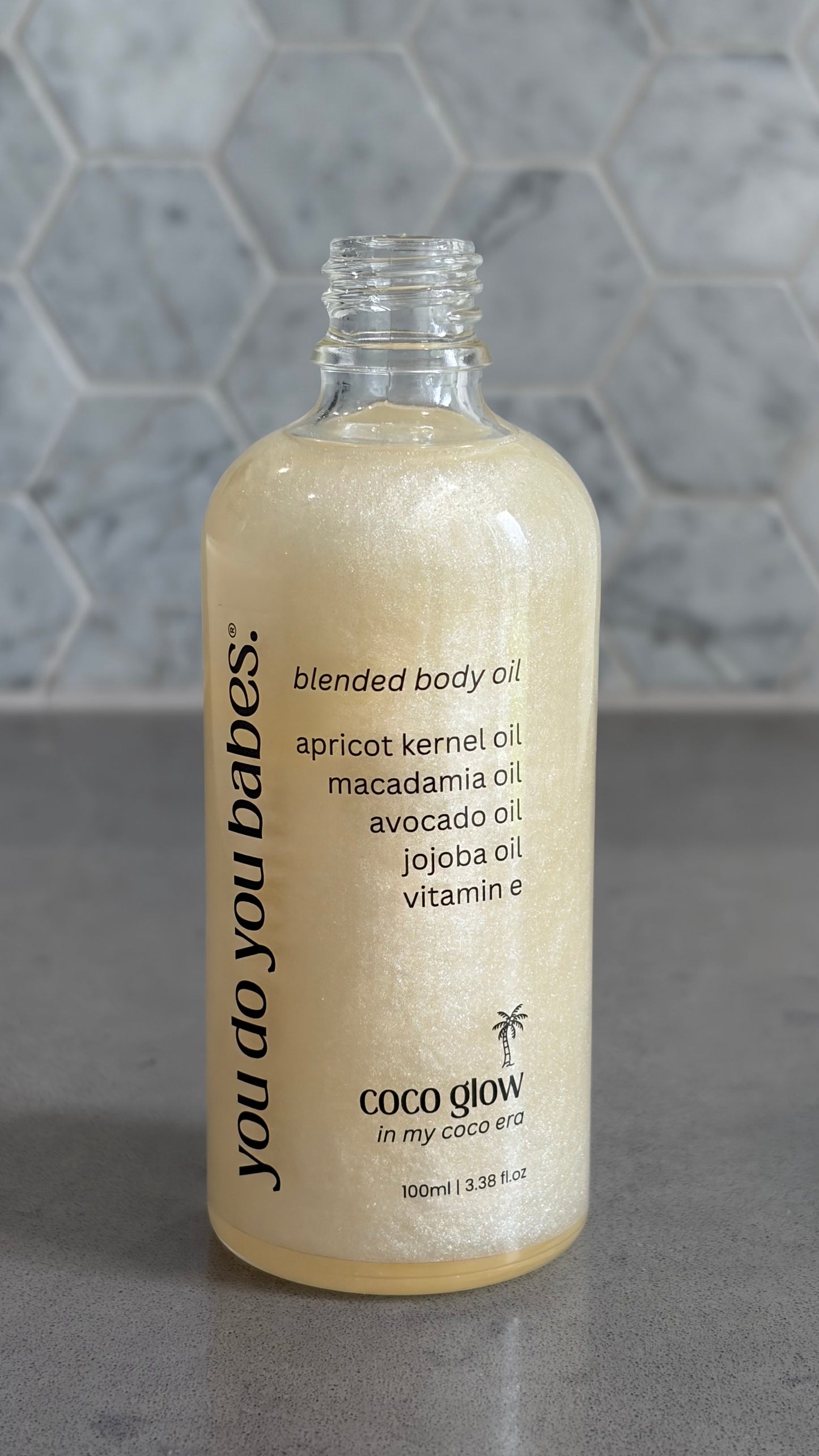 Bottle of 'you do you babes' coco glow blended body oil against a grey tiled wall.