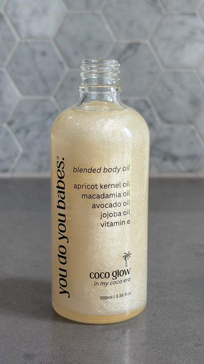 Bottle of 'you do you babes' coco glow blended body oil against a grey tiled wall.
