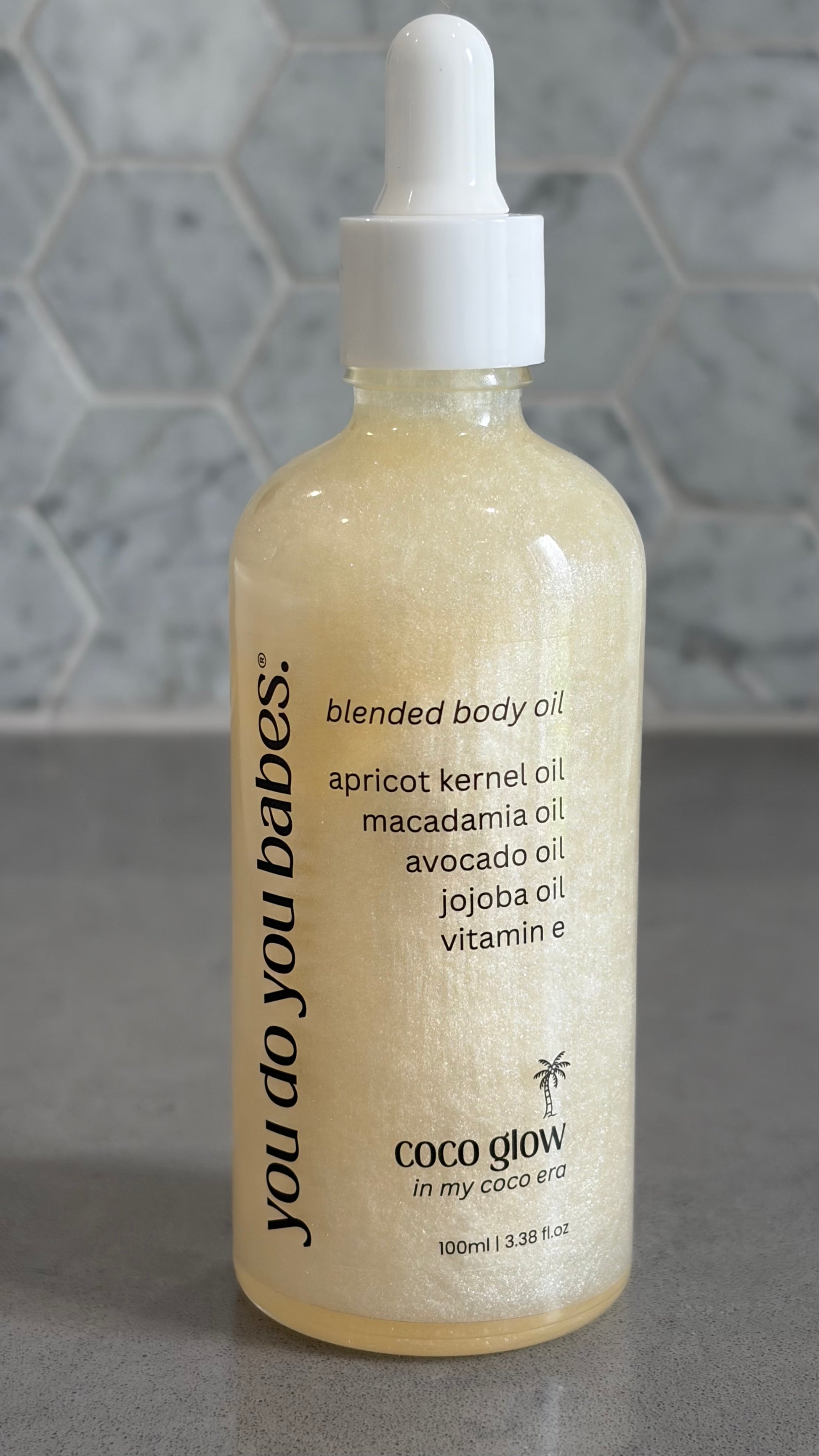 Bottle of 'you do you babes' coco glow blended body oil against a grey tiled wall.