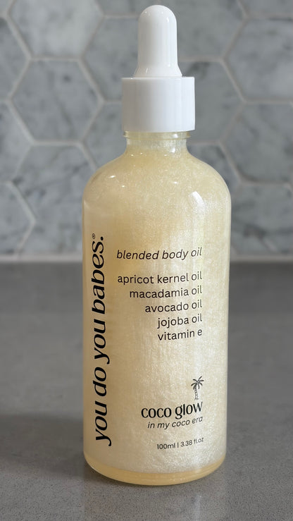 Bottle of 'you do you babes' coco glow blended body oil against a grey tiled wall.