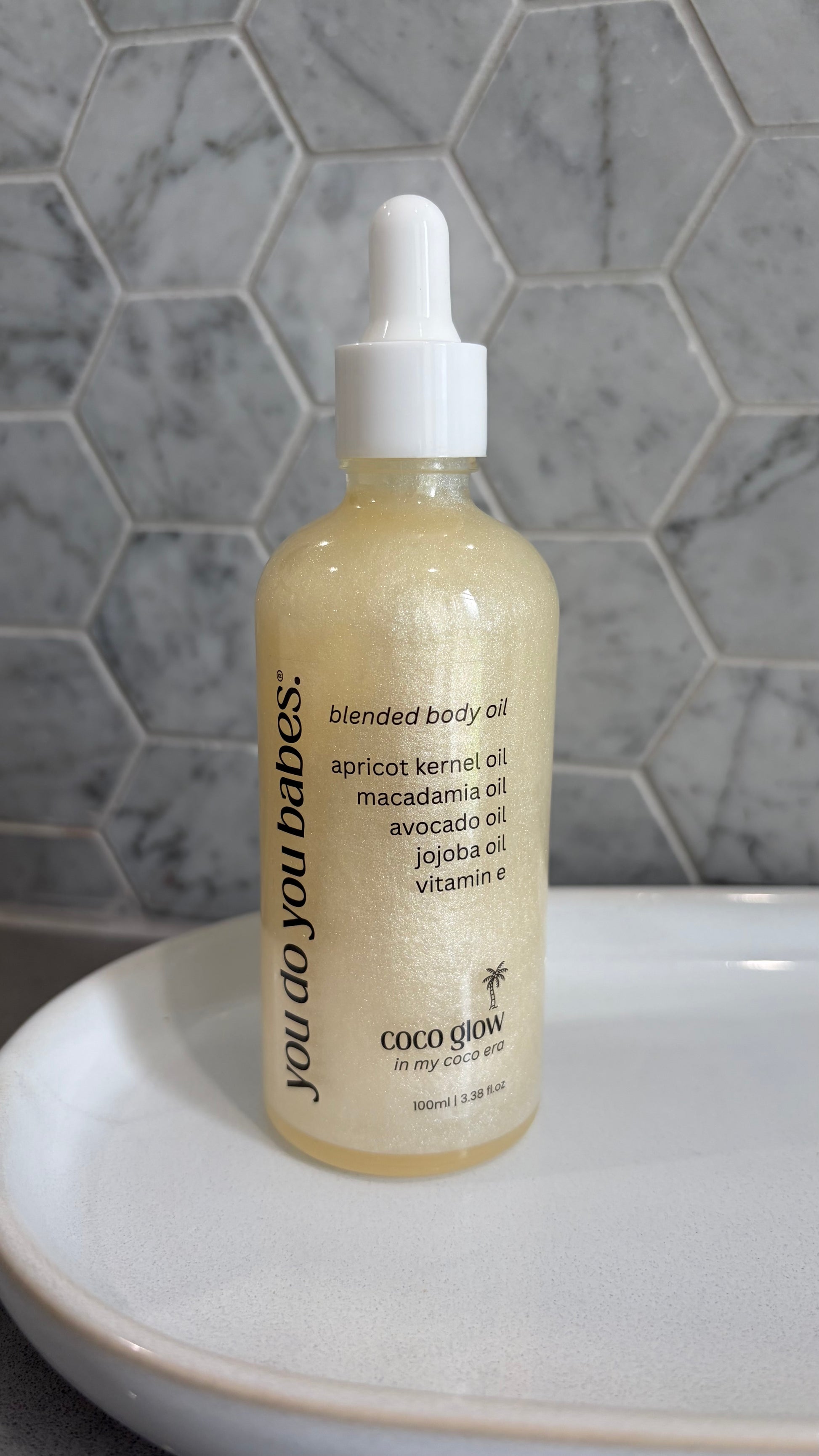 Bottle of 'you do you babes' coco glow blended body oil against a grey tiled wall.