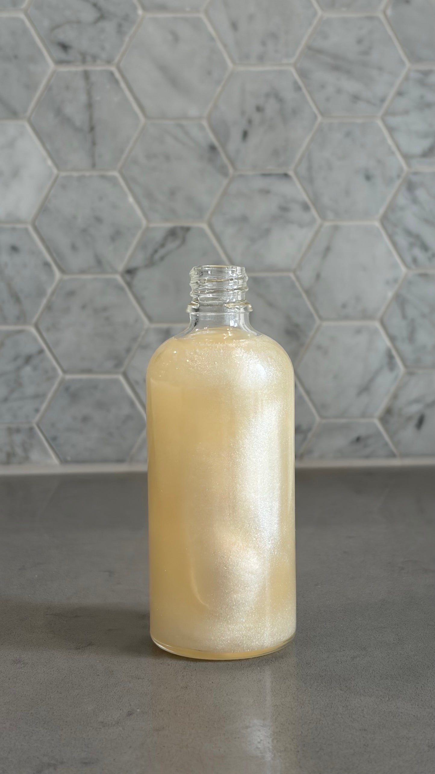 Bottle of 'you do you babes' coco glow blended body oil against a grey tiled wall.