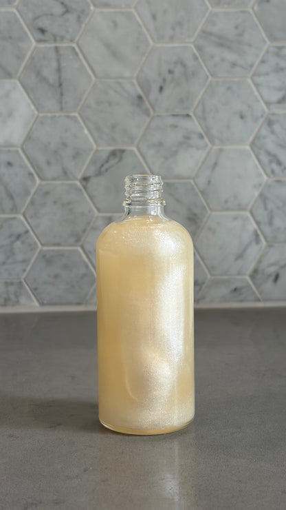 Bottle of 'you do you babes' coco glow blended body oil against a grey tiled wall.