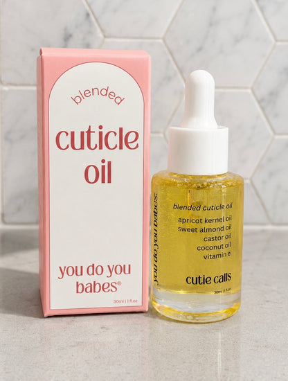 Bottle of cuticle oil with a pink box labeled 'you do you babes' on a light gray surface.