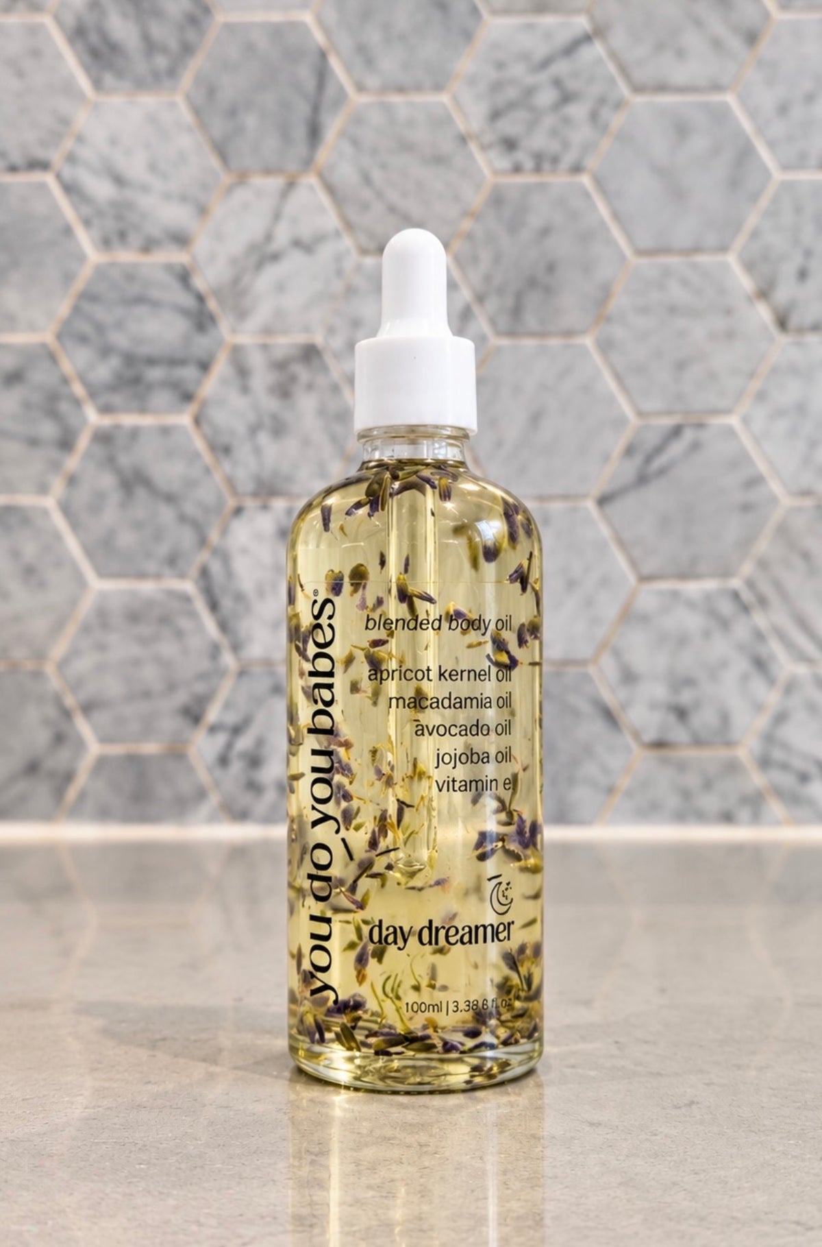 A 100ml dropper bottle of Day Dreamer 'You Do You Babes' blended body oil, filled with golden oil and dried botanicals, set against a gray hexagonal tile background.