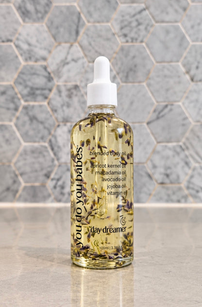 A 100ml dropper bottle of Day Dreamer 'You Do You Babes' blended body oil, filled with golden oil and dried botanicals, set against a gray hexagonal tile background.
