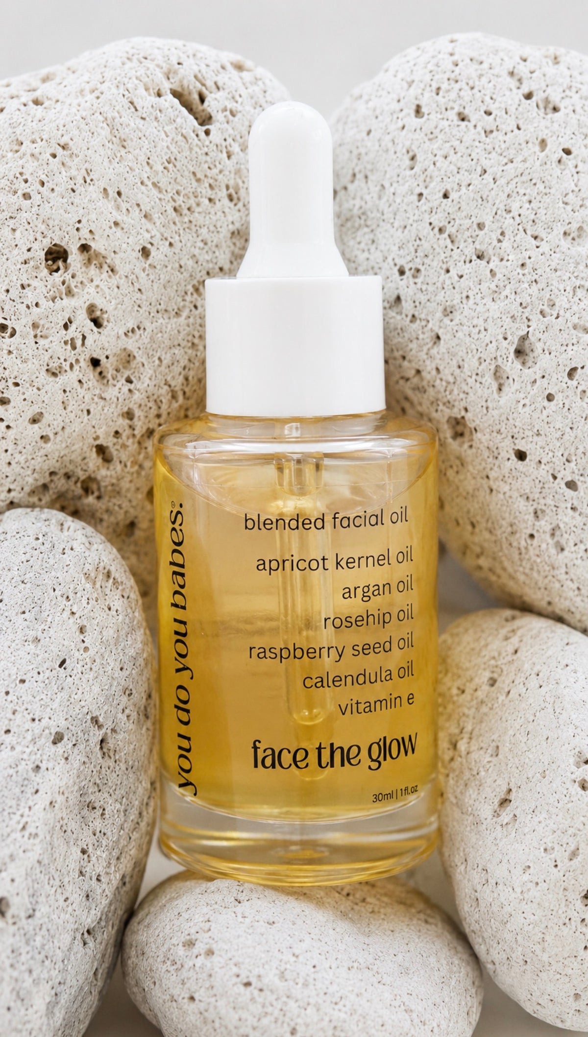 A clear glass dropper bottle of face the glow blended facial oil with a white dropper cap, surrounded by textured white volcanic stones. The oil inside is golden yellow, and the label lists ingredients like apricot kernel oil and argan oil.