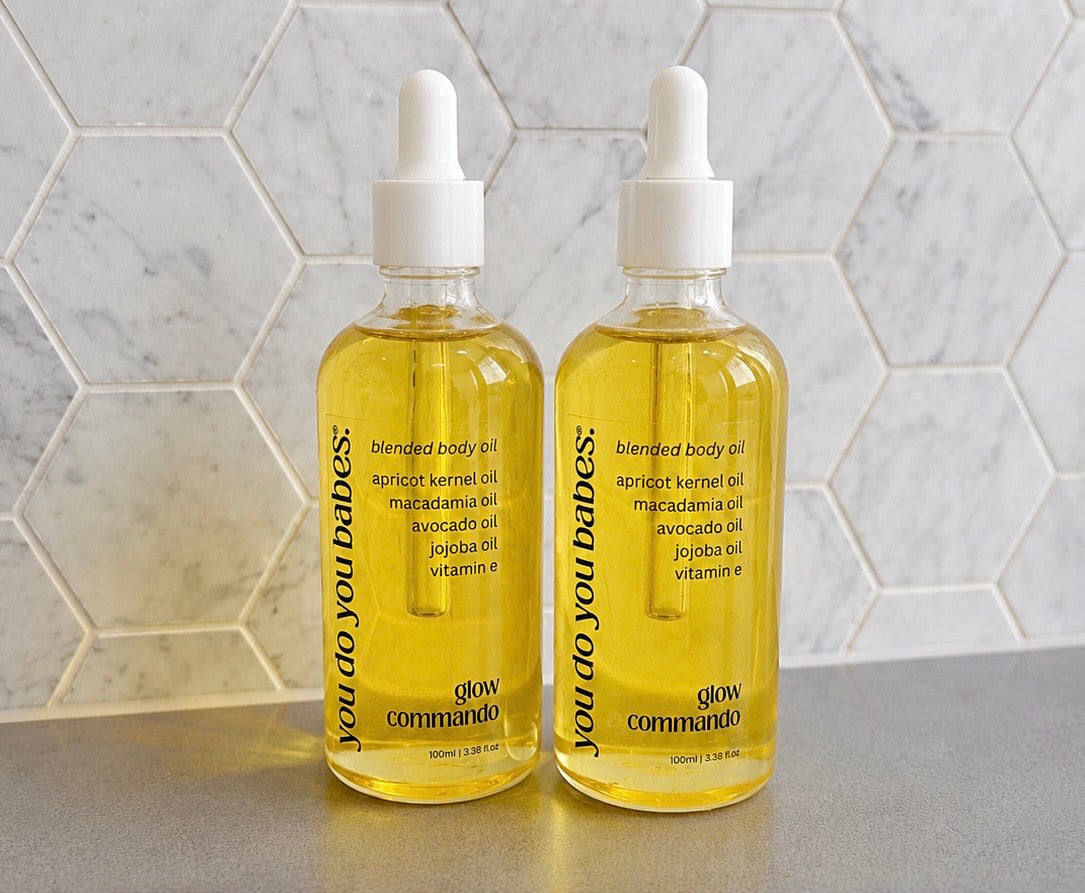 Two bottles of 'you do you babes' blended body oil on a marble countertop with a hexagonal tile backsplash.