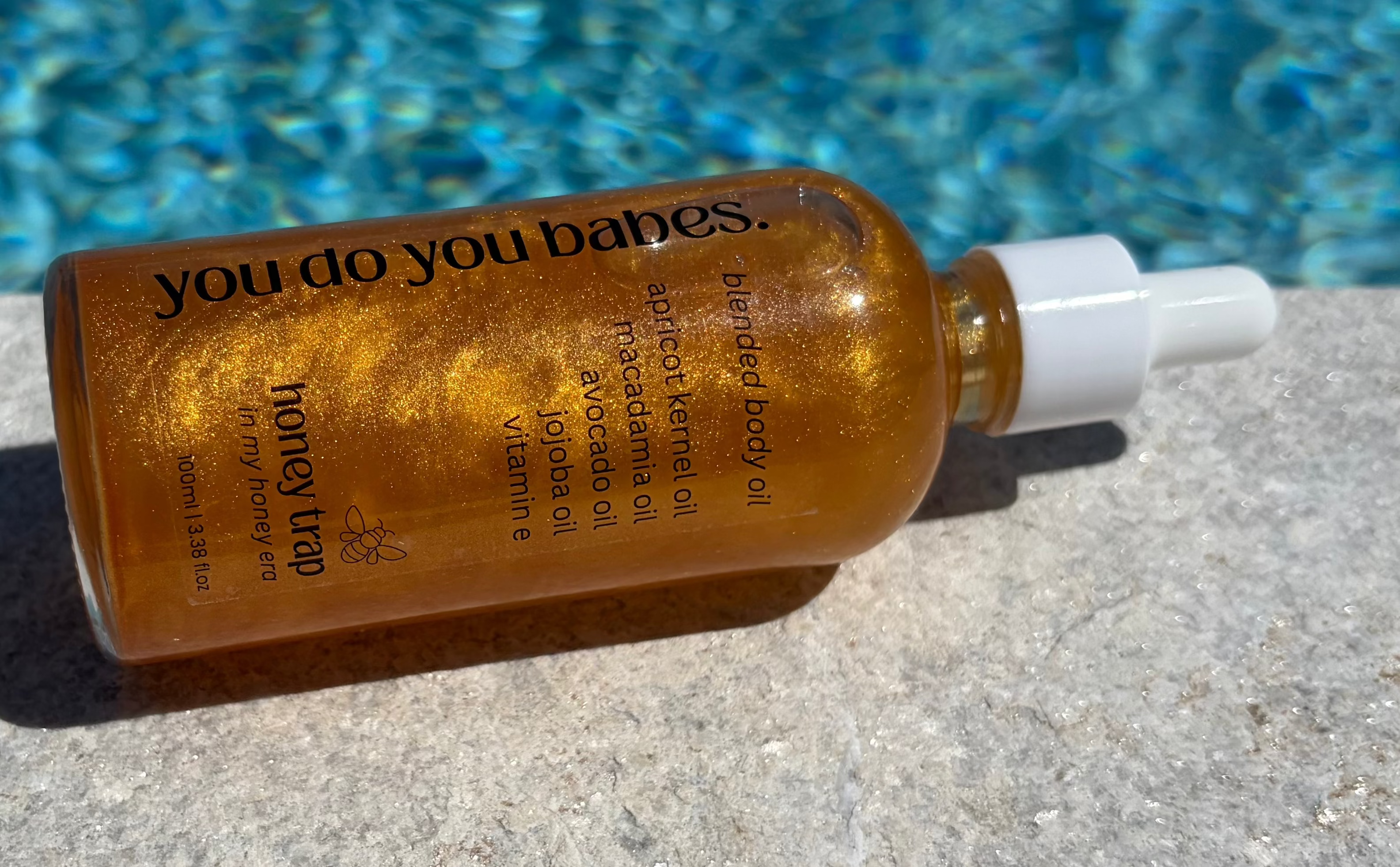 body oil bottle of honey trap from you do you babes with text on a concrete surface near a pool
