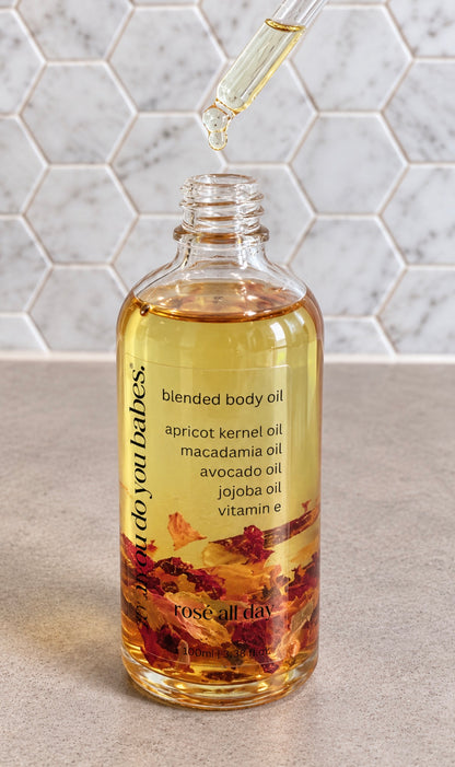 A clear glass bottle of rose all day blended body oil with a glass dropper above the open bottle, set against a light gray marble hexagonal tile background.