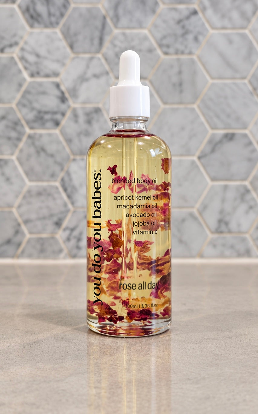 A clear glass dropper bottle of rose all over body butter with a white dropper cap, filled with golden-yellow oil and decorated with dried rose petals. The bottle is set on a light marble countertop against a matching hexagonal tile backsplash.