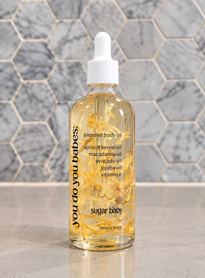 A clear bottle of 'you do you babes' blended body oil by sugar baby, filled with golden oil and suspended botanical pieces, featuring a white dropper cap, set against a gray marble hexagonal tile background.