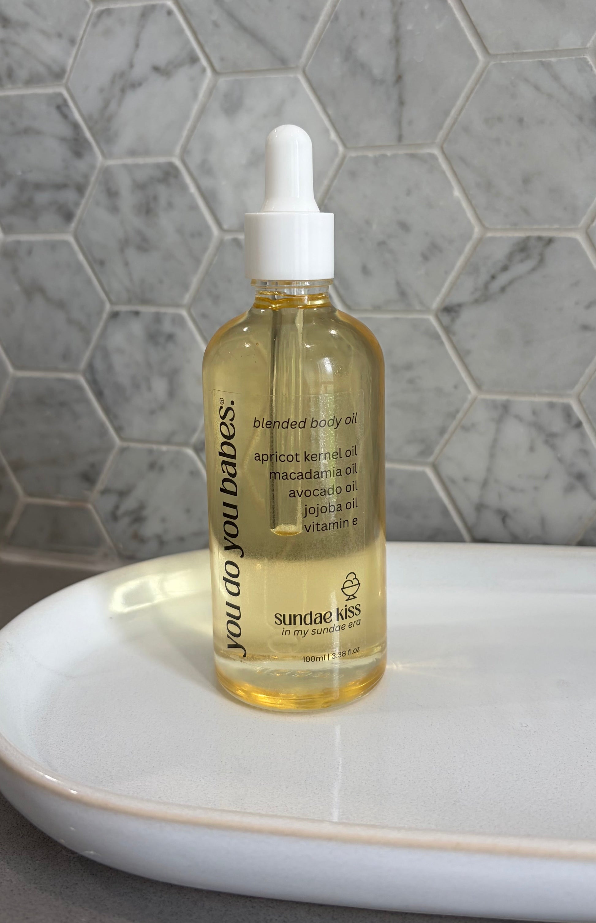 Bottle of sundae kiss body oil with a dropper on a white surface against a marble background