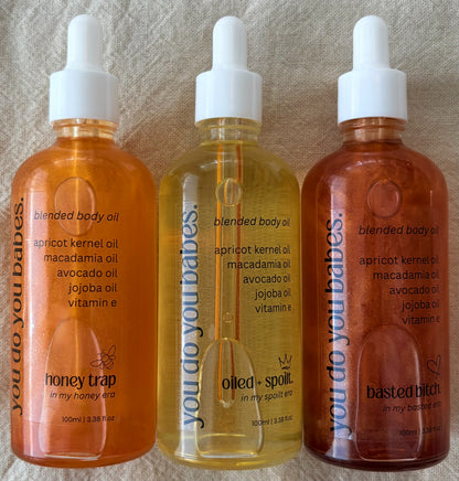 Three bottles of blended body oil from you do you babes with different names and descriptions on a textured background.