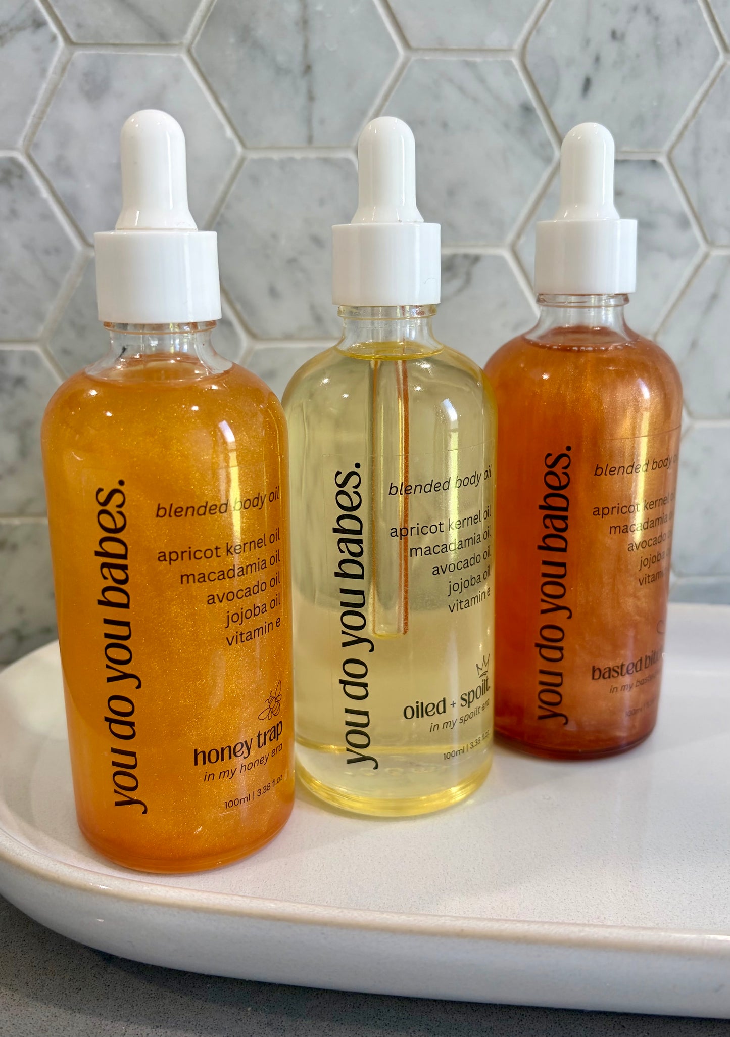 Three bottles of 'you do you babes' body oils on a white tray with a tiled wall background.