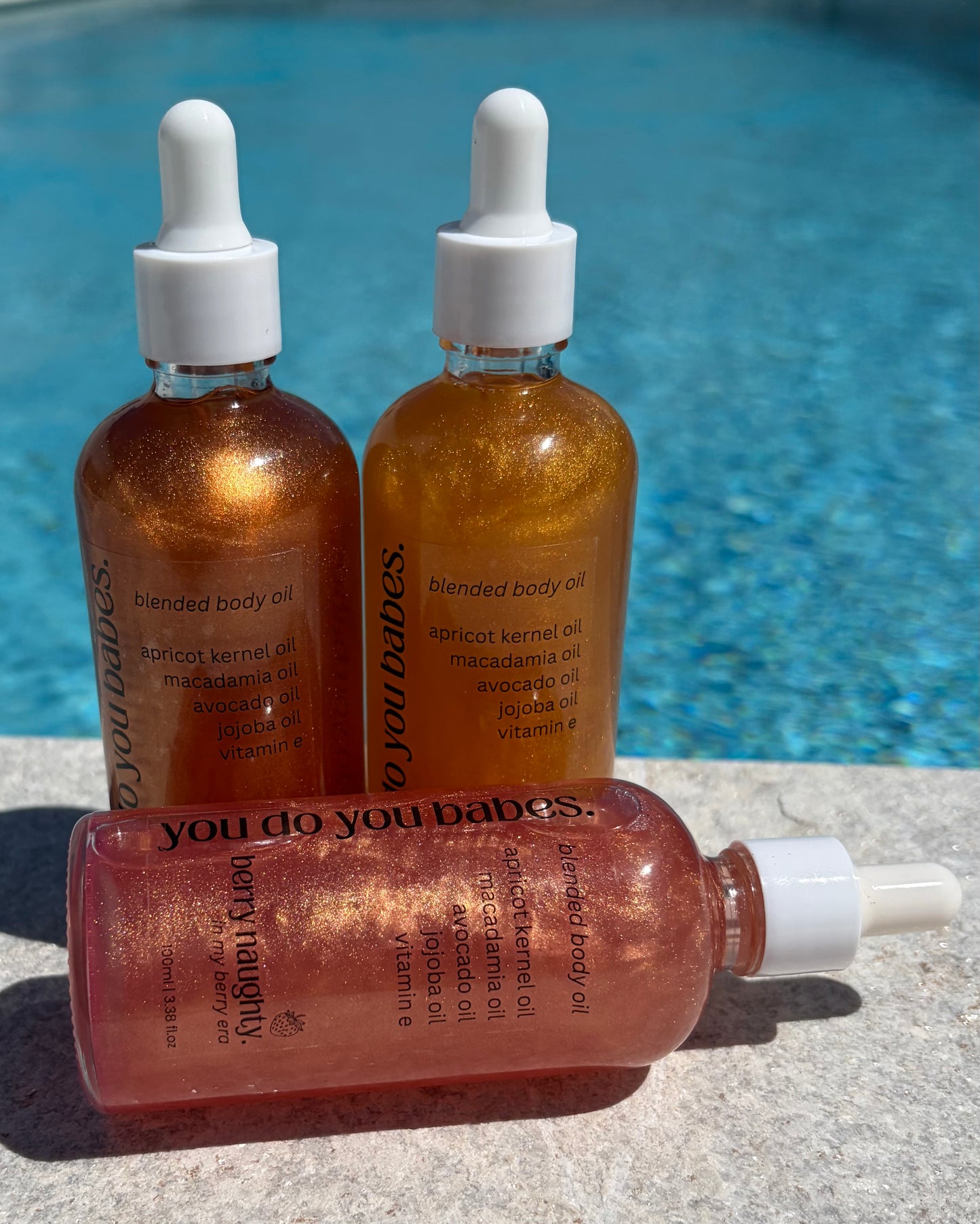 Three bottles of 'you do you babes' shimmer blended body oil with droppers against a pool background.