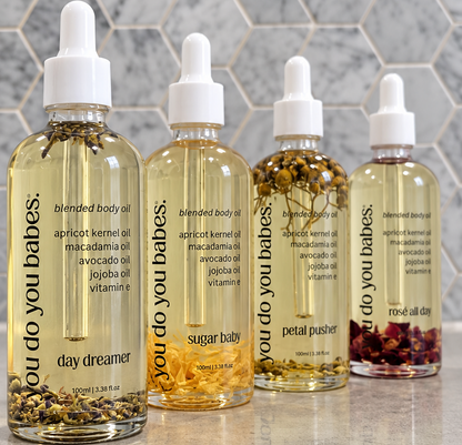 Four clear dropper bottles of body oil arranged on a glossy surface against a gray hexagonal tile wall. The bottles contain pale yellow oil with visible botanical pieces at the bottom, and the labels read 'you do you babes.